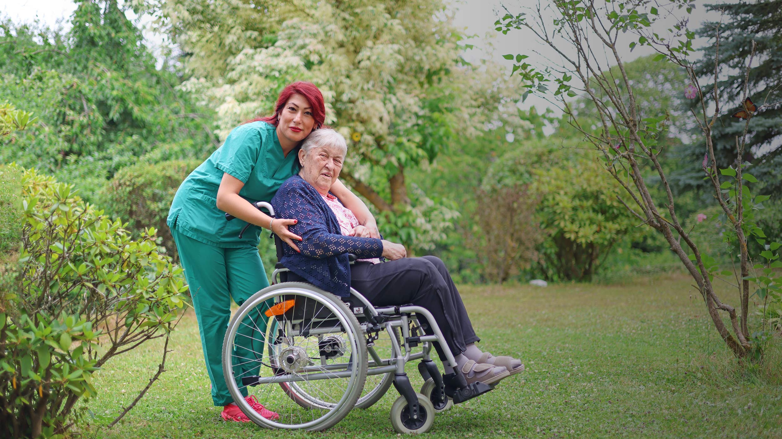 Eine Plegefachkraft und eine Seniorin im Rollstuhl in einem blühenden Garten.